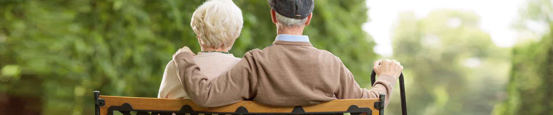 Senior man with cane and his wife take a break on a park bench; view of their backs as they face away.